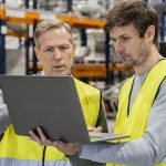 Dois homens de coletes amarelos olhando para tela do notebook dentro da fabrica
