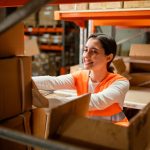 imagem exibe uma mulher sorridente, também com um colete de segurança laranja, organizando caixas em um armazém. Ela está em um ambiente de trabalho, dentro de um depósito, com prateleiras de caixas ao fundo. A expressão dela é alegre e focada na tarefa, transmitindo um senso de produtividade e otimismo no ambiente de trabalho.