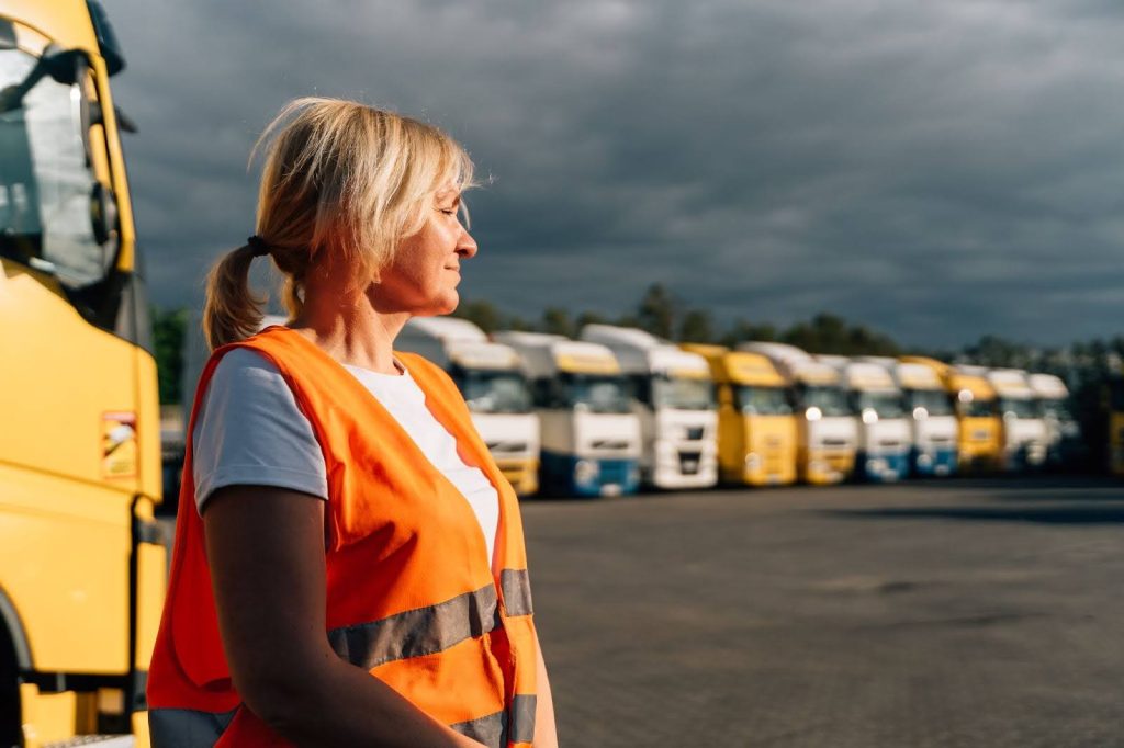 imagem mostra uma mulher de colete de segurança laranja, com cabelos loiros, observando de perfil para um grupo de caminhões estacionados ao fundo. Ela está em um ambiente aberto, possivelmente em um pátio de transporte, e o céu ao fundo apresenta nuvens escuras, sugerindo um clima de mudança.