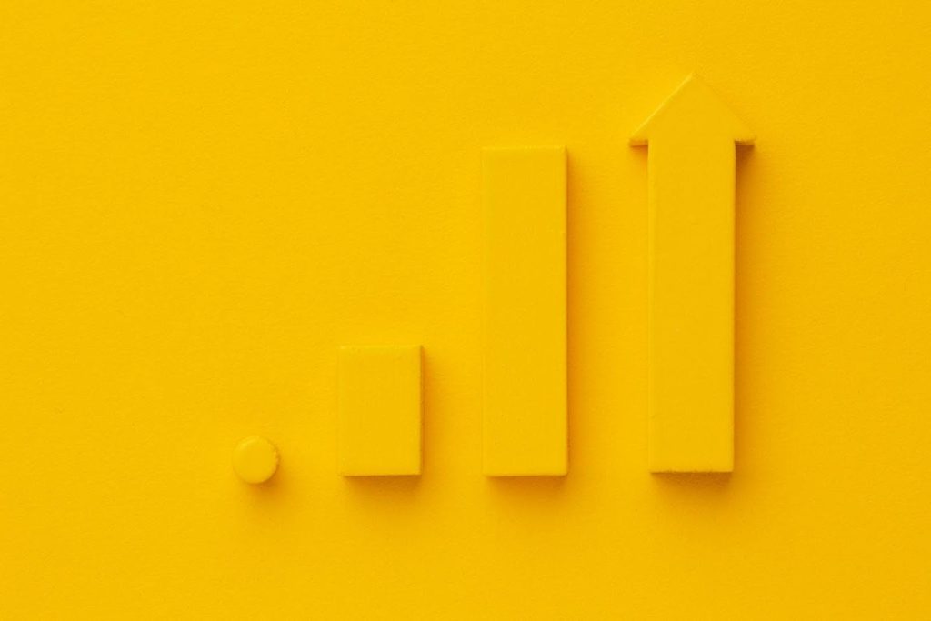 A imagem é composta por blocos e uma seta, todos de cor amarela, organizados em uma formação que lembra um gráfico de barras. O fundo também é amarelo, fazendo com que os elementos se destaquem. Os blocos estão alinhados de forma crescente, com a seta no final apontando para cima, sugerindo progresso ou aumento.
