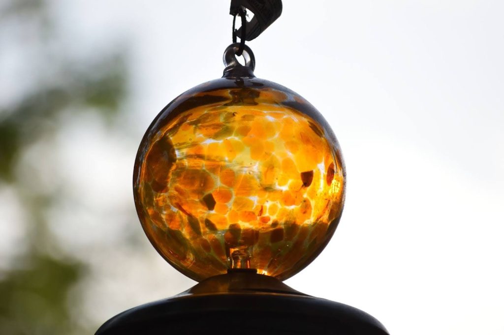 A imagem mostra uma esfera de vidro âmbar, pendurada, com luz natural ao fundo. A esfera reflete a luz e mostra um padrão de cores quentes e suaves, criando um efeito visual translúcido. Ela está suspensa, com um contraste suave do fundo mais claro.