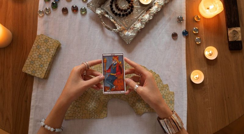 A imagem apresenta uma mesa com uma toalha clara, onde estão espalhados diversos itens místicos. Uma pessoa está segurando uma carta de tarô, representando 
